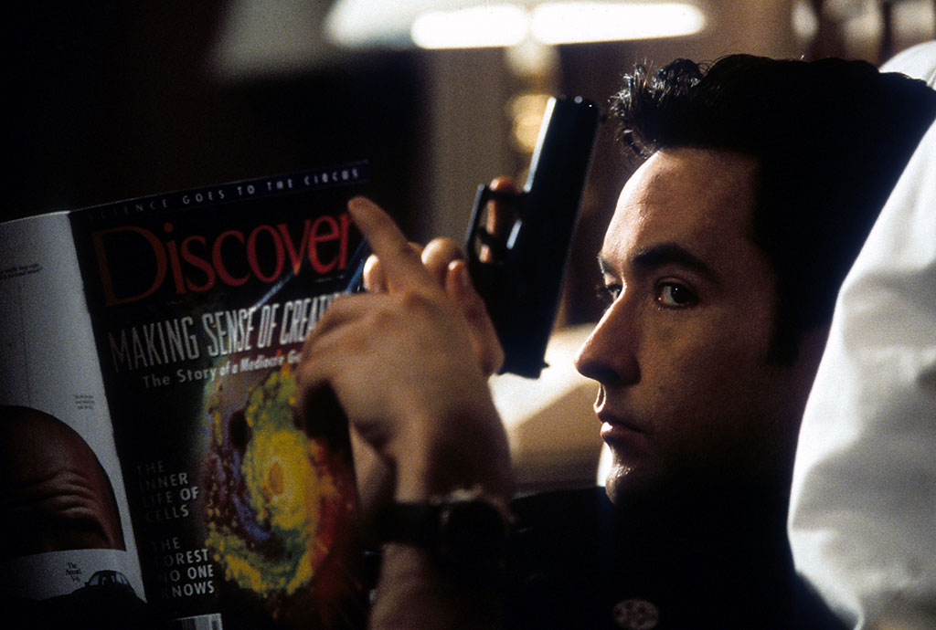 John Cusack holding a gun and reading.